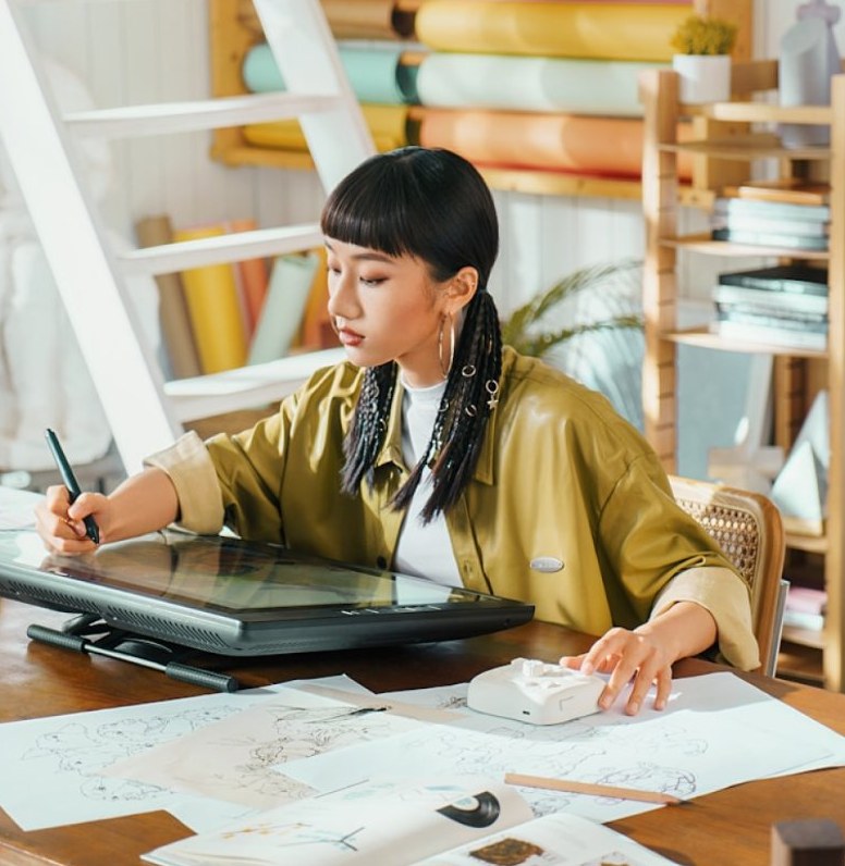 Zoe sketching on a pen display, surrounded by paper drawings and bookshelves